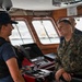 The image shows a conversation on the bridge of a U.S. Coast Guard vessel between a female Coast Guard member in tactical vest and a male U.S. Marine Corps officer in MARPAT camouflage uniform. A third military personnel member is visible in the background. The setting appears to be a ship's bridge or command center with navigation equipment visible.