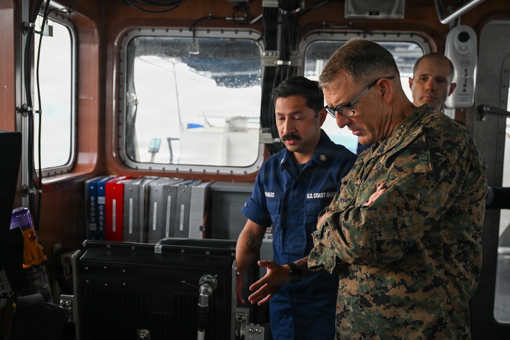 The image shows the interior of a U.S. Coast Guard vessel bridge where a Coast Guard member is briefing a U.S. Marine Corps officer in camouflage uniform. The Coast Guard member, whose name tag reads 'MORALES' and whose uniform is labeled 'U.S. COAST GUARD,' appears to be explaining equipment or operations at the helm console. A third military individual is visible in the background.