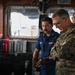 The image shows the interior of a U.S. Coast Guard vessel bridge where a Coast Guard member is briefing a U.S. Marine Corps officer in camouflage uniform. The Coast Guard member, whose name tag reads 'MORALES' and whose uniform is labeled 'U.S. COAST GUARD,' appears to be explaining equipment or operations at the helm console. A third military individual is visible in the background.
