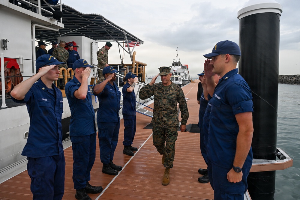 A U.S. Marine Corps officer in woodland MARPAT camouflage uniform is walking along a dock while being rendered honors by a side boy formation of U.S. Coast Guard personnel in blue operational dress uniforms. The ceremony appears to be a formal boarding or departure ritual conducted on a floating pier adjacent to a Coast Guard vessel. The setting is an overcast maritime environment with additional military personnel visible on the vessel's deck.