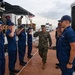 A U.S. Marine Corps officer in woodland MARPAT camouflage uniform is walking along a dock while being rendered honors by a side boy formation of U.S. Coast Guard personnel in blue operational dress uniforms. The ceremony appears to be a formal boarding or departure ritual conducted on a floating pier adjacent to a Coast Guard vessel. The setting is an overcast maritime environment with additional military personnel visible on the vessel's deck.