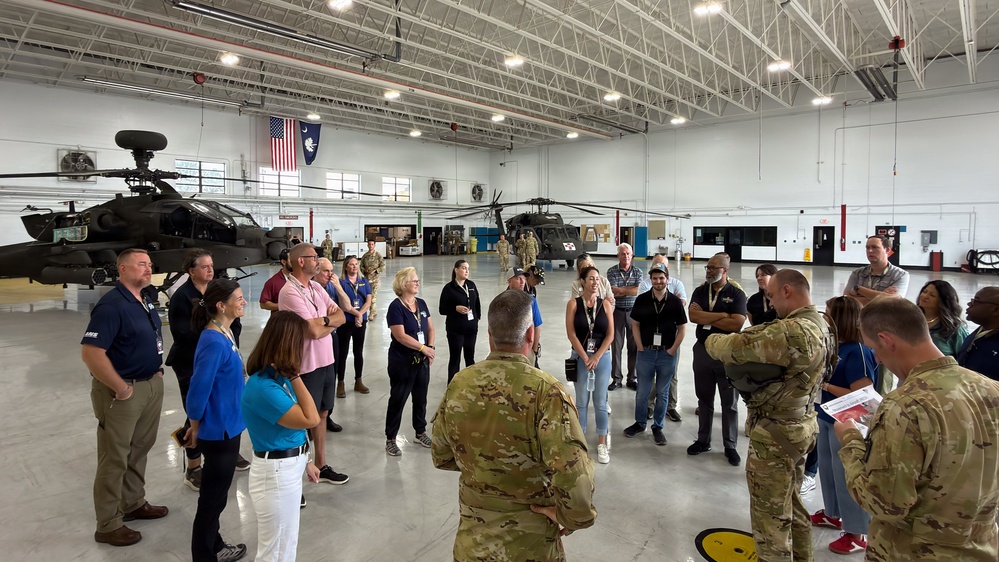 A group of civilians wearing visitor badges are gathered in a military helicopter hangar for what appears to be an official briefing or tour. Two military personnel in Army Combat Uniforms (ACU/OCP pattern) are addressing the group. The hangar contains at least two military helicopters, including an AH-64 Apache attack helicopter and a UH-60 Black Hawk utility helicopter.