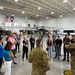A group of civilians wearing visitor badges are gathered in a military helicopter hangar for what appears to be an official briefing or tour. Two military personnel in Army Combat Uniforms (ACU/OCP pattern) are addressing the group. The hangar contains at least two military helicopters, including an AH-64 Apache attack helicopter and a UH-60 Black Hawk utility helicopter.