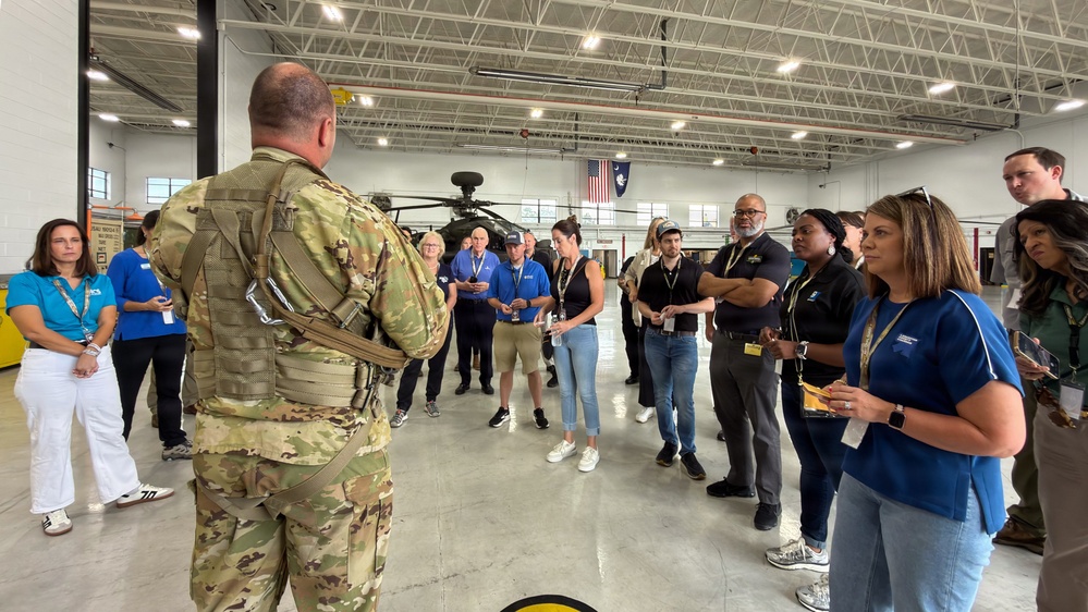 A military personnel in full combat gear and tactical vest is briefing a group of civilians inside a large hangar facility. The civilians, many wearing blue shirts and lanyards with badges, appear to be on an organized tour or employer support program visit. An Apache attack helicopter is visible in the background along with American and state flags.