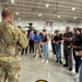 A military personnel in full combat gear and tactical vest is briefing a group of civilians inside a large hangar facility. The civilians, many wearing blue shirts and lanyards with badges, appear to be on an organized tour or employer support program visit. An Apache attack helicopter is visible in the background along with American and state flags.