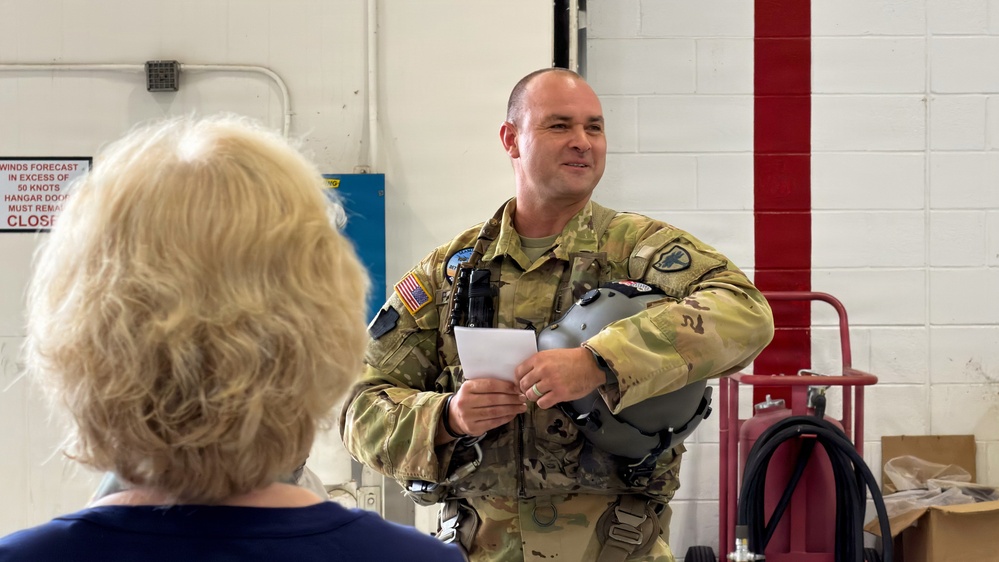 A U.S. military service member in Army Combat Uniform (ACU) with flight gear stands inside what appears to be a hangar facility, holding a flight helmet and a small card or paper. He appears to be speaking or presenting to a blonde-haired civilian woman whose back is to the camera. The setting suggests a briefing or orientation event at a military aviation facility.