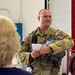 A U.S. military service member in Army Combat Uniform (ACU) with flight gear stands inside what appears to be a hangar facility, holding a flight helmet and a small card or paper. He appears to be speaking or presenting to a blonde-haired civilian woman whose back is to the camera. The setting suggests a briefing or orientation event at a military aviation facility.