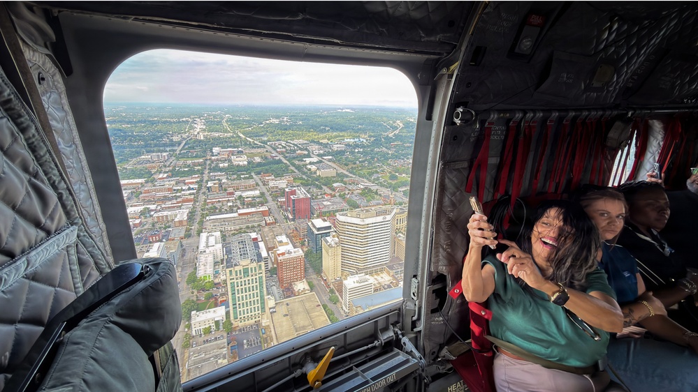 Passengers are seated inside a large military-style helicopter, viewing and photographing an aerial cityscape through the open rear cargo door. A woman in a green top is smiling and holding up a phone to capture the view below. The city visible through the opening features a downtown urban core with high-rise buildings surrounded by residential and commercial neighborhoods.