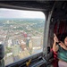 Passengers are seated inside a large military-style helicopter, viewing and photographing an aerial cityscape through the open rear cargo door. A woman in a green top is smiling and holding up a phone to capture the view below. The city visible through the opening features a downtown urban core with high-rise buildings surrounded by residential and commercial neighborhoods.