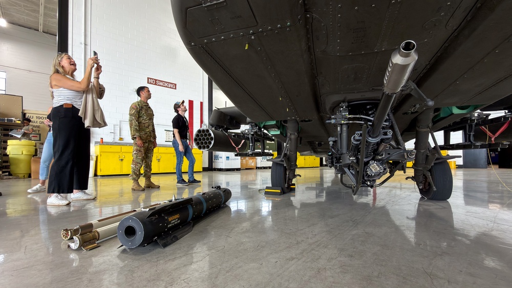 The image shows civilians and a military personnel inside an aircraft hangar, examining what appears to be a military helicopter from below. Several munitions, likely AGM-114 Hellfire missiles, are displayed on the hangar floor in the foreground. This appears to be an official military open house, media day, or public affairs event.