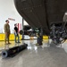 The image shows civilians and a military personnel inside an aircraft hangar, examining what appears to be a military helicopter from below. Several munitions, likely AGM-114 Hellfire missiles, are displayed on the hangar floor in the foreground. This appears to be an official military open house, media day, or public affairs event.
