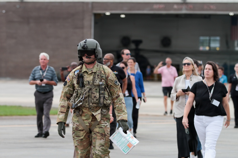 A U.S. Army soldier in full flight gear, including a helmet with visor and camouflage uniform, walks across what appears to be a military airfield or tarmac. The soldier is leading or walking ahead of a group of civilians, suggesting this may be a public affairs event, base tour, or open house. A military hangar is visible in the background with what appears to be a helicopter inside.