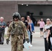 A U.S. Army soldier in full flight gear, including a helmet with visor and camouflage uniform, walks across what appears to be a military airfield or tarmac. The soldier is leading or walking ahead of a group of civilians, suggesting this may be a public affairs event, base tour, or open house. A military hangar is visible in the background with what appears to be a helicopter inside.