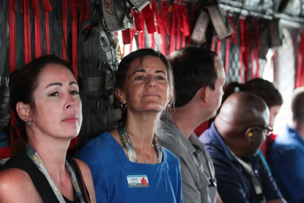 A group of civilians are seated inside what appears to be a military transport aircraft, identifiable by the red safety straps, cargo netting, and equipment visible along the fuselage walls. Several individuals are wearing lanyards with 'Army National Guard' branding, and one woman in the center is wearing a name badge reading 'Laura'. The group appears to be on an orientation or familiarization flight.