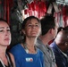 A group of civilians are seated inside what appears to be a military transport aircraft, identifiable by the red safety straps, cargo netting, and equipment visible along the fuselage walls. Several individuals are wearing lanyards with 'Army National Guard' branding, and one woman in the center is wearing a name badge reading 'Laura'. The group appears to be on an orientation or familiarization flight.
