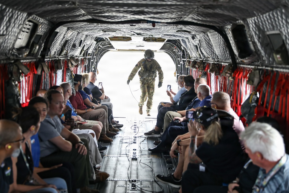 The image shows the interior of a military transport helicopter, likely a CH-47 Chinook, with civilians seated along both sides of the cargo bay. A uniformed soldier in full combat gear and helmet stands at the open rear ramp, facing the seated passengers. The civilians appear to be on an official tour or demonstration, with several photographing the soldier and surroundings.