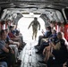 The image shows the interior of a military transport helicopter, likely a CH-47 Chinook, with civilians seated along both sides of the cargo bay. A uniformed soldier in full combat gear and helmet stands at the open rear ramp, facing the seated passengers. The civilians appear to be on an official tour or demonstration, with several photographing the soldier and surroundings.