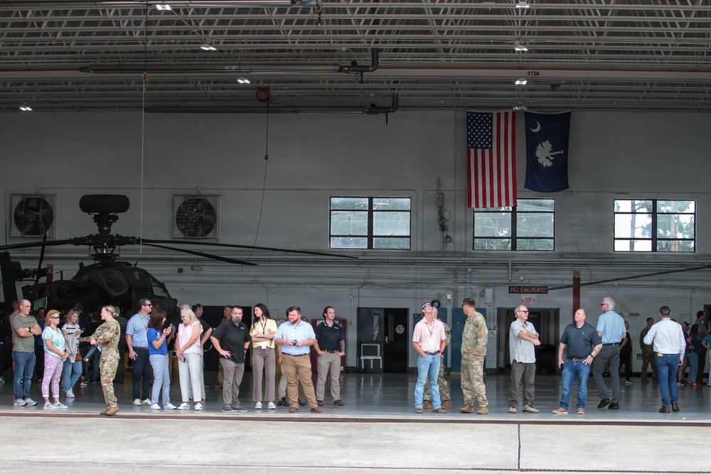 A group of civilians and military personnel are gathered inside a large military helicopter hangar, likely during an official visit or tour. The hangar contains at least one military helicopter, possibly an AH-64 Apache, and displays both the United States flag and the South Carolina state flag. The setting suggests this is a National Guard or Army installation in South Carolina.