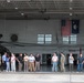 A group of civilians and military personnel are gathered inside a large military helicopter hangar, likely during an official visit or tour. The hangar contains at least one military helicopter, possibly an AH-64 Apache, and displays both the United States flag and the South Carolina state flag. The setting suggests this is a National Guard or Army installation in South Carolina.