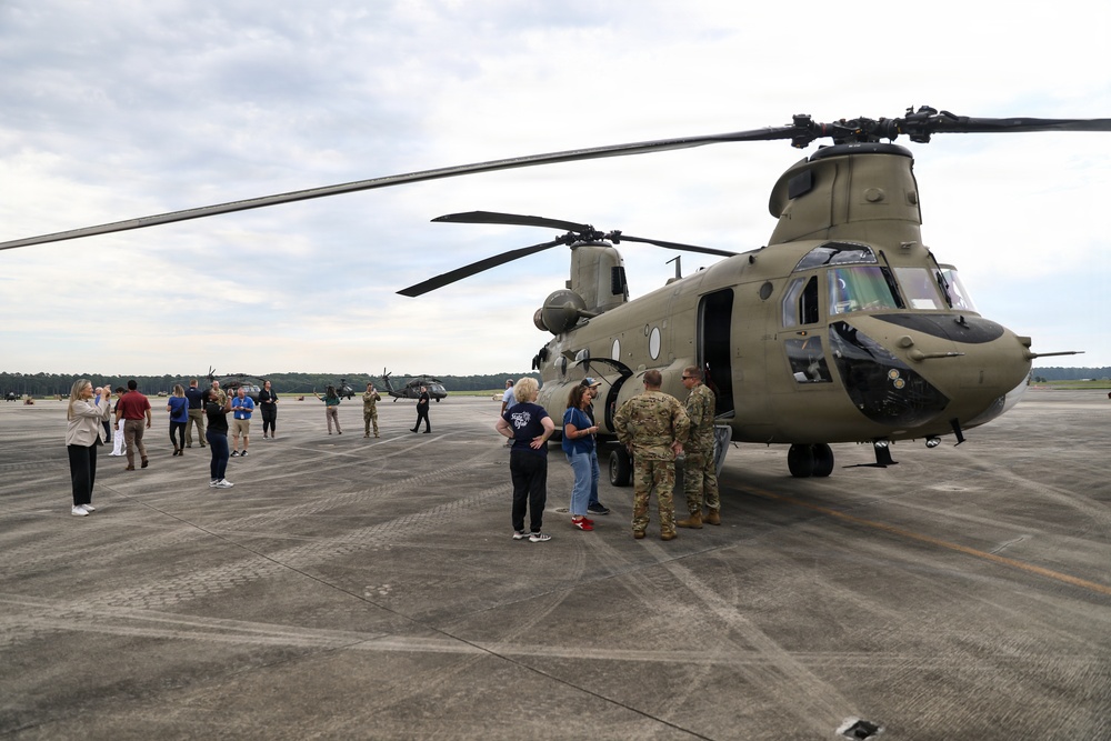 A group of civilians and military personnel are gathered on a tarmac around a U.S. Army CH-47 Chinook tandem-rotor helicopter during what appears to be a public outreach or familiarization event. Military personnel in camouflage uniforms are speaking with civilians near the open forward door of the aircraft. Additional helicopters, including what appear to be Black Hawks, are visible in the background.