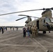 A group of civilians and military personnel are gathered on a tarmac around a U.S. Army CH-47 Chinook tandem-rotor helicopter during what appears to be a public outreach or familiarization event. Military personnel in camouflage uniforms are speaking with civilians near the open forward door of the aircraft. Additional helicopters, including what appear to be Black Hawks, are visible in the background.
