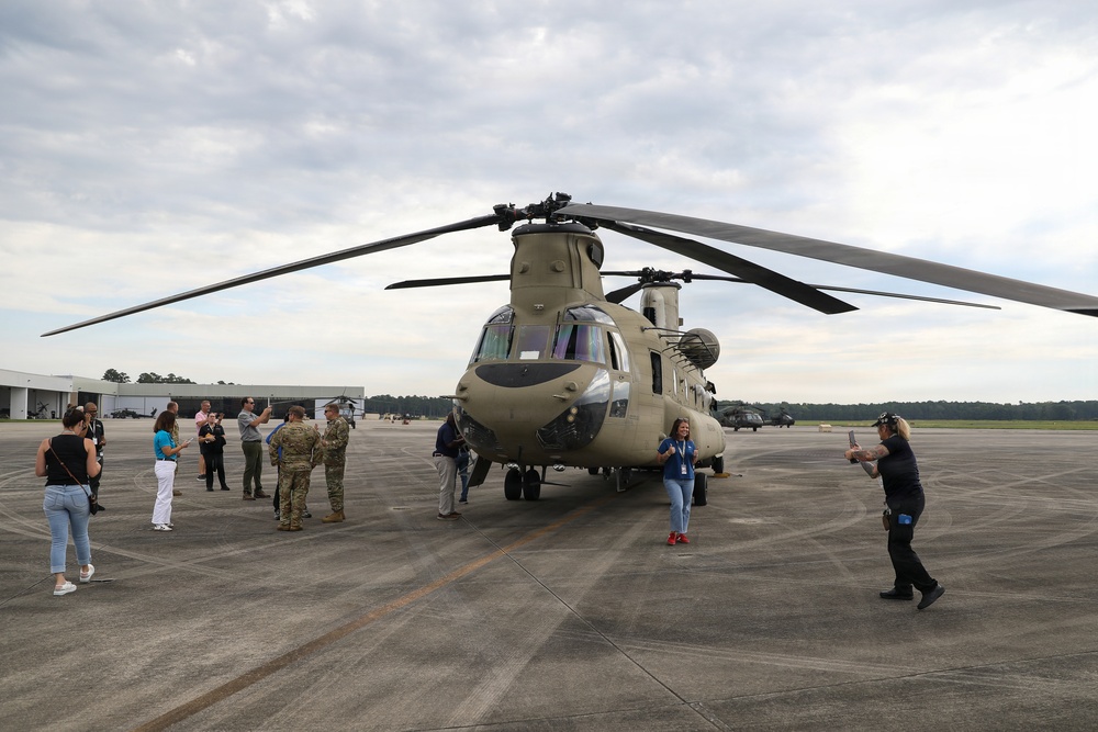 A large military tandem-rotor helicopter, identifiable as a Boeing CH-47 Chinook in olive drab livery, is parked on a tarmac at what appears to be a military airfield. A group of civilians and uniformed military personnel are gathered around the aircraft, suggesting a public affairs event, media day, or community outreach visit. Several individuals are photographing the helicopter and posing near it.