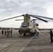 A large military tandem-rotor helicopter, identifiable as a Boeing CH-47 Chinook in olive drab livery, is parked on a tarmac at what appears to be a military airfield. A group of civilians and uniformed military personnel are gathered around the aircraft, suggesting a public affairs event, media day, or community outreach visit. Several individuals are photographing the helicopter and posing near it.