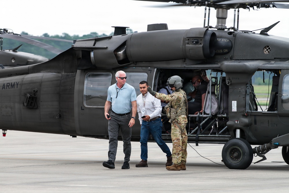 The image shows a group of individuals near a U.S. Army UH-60 Black Hawk helicopter on a tarmac or airfield. A uniformed soldier in multicam/OCP pattern is interacting with two civilians, while additional personnel are visible inside the open cabin of the helicopter. A second Army helicopter is partially visible in the background.