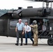 The image shows a group of individuals near a U.S. Army UH-60 Black Hawk helicopter on a tarmac or airfield. A uniformed soldier in multicam/OCP pattern is interacting with two civilians, while additional personnel are visible inside the open cabin of the helicopter. A second Army helicopter is partially visible in the background.