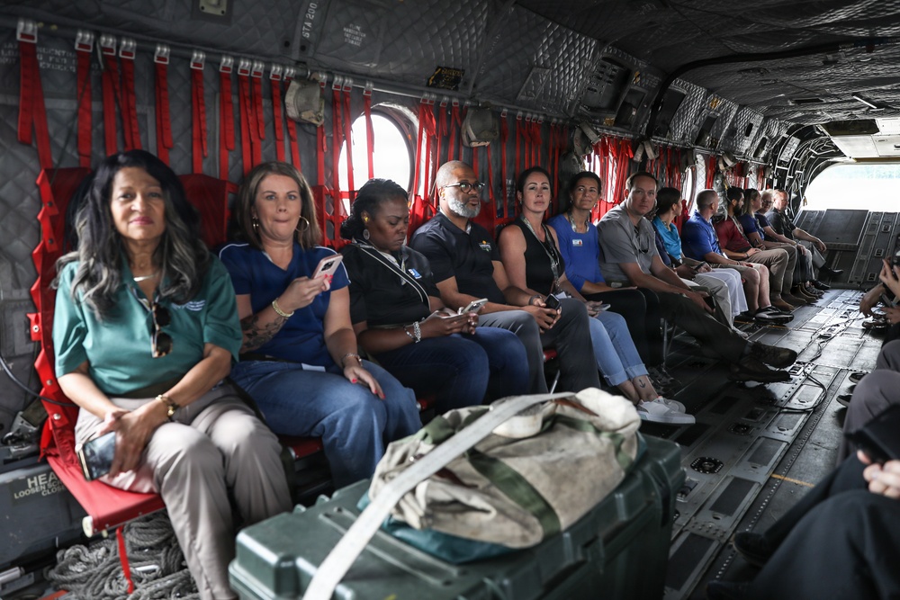 A group of approximately 15 civilians are seated along the interior walls of a military transport helicopter, likely a CH-47 Chinook, based on the distinctive interior configuration. The passengers appear to be on an official tour or briefing flight, dressed in casual civilian attire. Red cargo straps, military equipment bags, and safety markings are visible throughout the aircraft interior.
