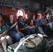 A group of approximately 15 civilians are seated along the interior walls of a military transport helicopter, likely a CH-47 Chinook, based on the distinctive interior configuration. The passengers appear to be on an official tour or briefing flight, dressed in casual civilian attire. Red cargo straps, military equipment bags, and safety markings are visible throughout the aircraft interior.