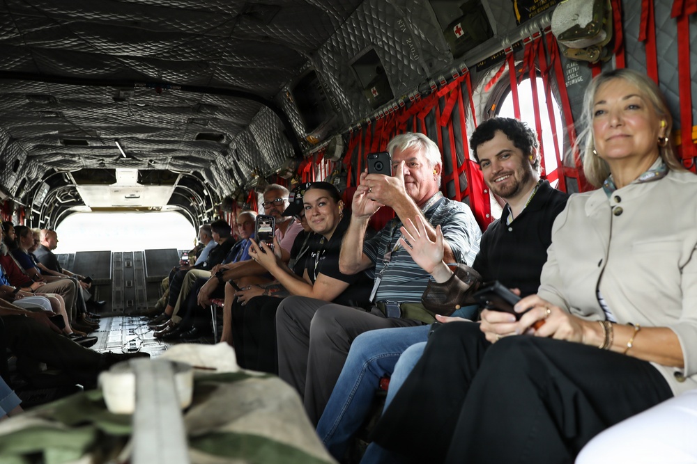 A group of civilians are seated inside the cargo bay of a military helicopter, likely a CH-47 Chinook, based on the distinctive interior features including red cargo straps and diamond-pattern walls. Several passengers are using smartphones and cameras to document the experience. The open rear ramp is visible in the background, suggesting the aircraft may be on the ground or in flight.