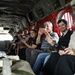 A group of civilians are seated inside the cargo bay of a military helicopter, likely a CH-47 Chinook, based on the distinctive interior features including red cargo straps and diamond-pattern walls. Several passengers are using smartphones and cameras to document the experience. The open rear ramp is visible in the background, suggesting the aircraft may be on the ground or in flight.