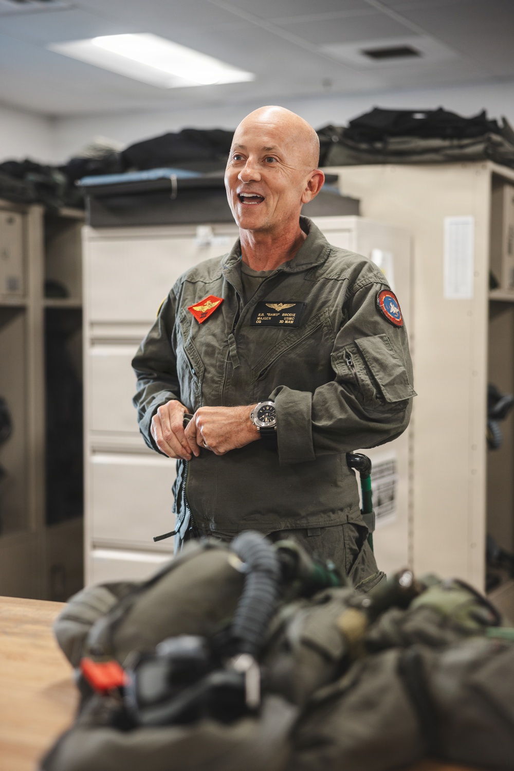 A senior military officer in a green flight suit stands in what appears to be a flight equipment room or locker room, smiling and engaged in conversation. His name tag and insignia indicate he is a Major General (CG) in the USMC assigned to the 3rd Marine Aircraft Wing (3D MAW). Flight gear and equipment are visible in the foreground on a table.