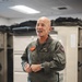 A senior military officer in a green flight suit stands in what appears to be a flight equipment room or locker room, smiling and engaged in conversation. His name tag and insignia indicate he is a Major General (CG) in the USMC assigned to the 3rd Marine Aircraft Wing (3D MAW). Flight gear and equipment are visible in the foreground on a table.