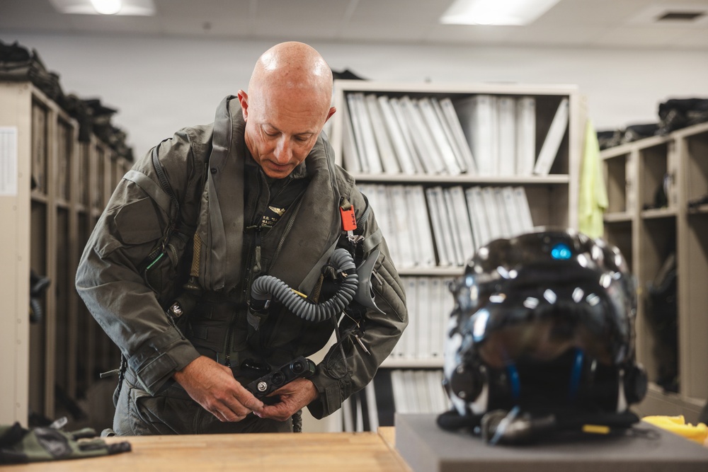 A bald male aviator in a military flight suit is preparing his gear in what appears to be a flight equipment room or locker room. He is adjusting or fastening equipment on his torso while a dark-colored advanced flight helmet sits on the table beside him. The setting includes storage shelving with binders and equipment lockers in the background.