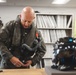 A bald male aviator in a military flight suit is preparing his gear in what appears to be a flight equipment room or locker room. He is adjusting or fastening equipment on his torso while a dark-colored advanced flight helmet sits on the table beside him. The setting includes storage shelving with binders and equipment lockers in the background.