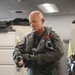 A bald middle-aged man wearing a military flight suit and tactical gear is standing in what appears to be a flight equipment room or locker room. He is donning fingerless tactical gloves and has an oxygen mask with corrugated hose draped over his shoulder. The setting suggests pre-flight preparation in a military aviation context.