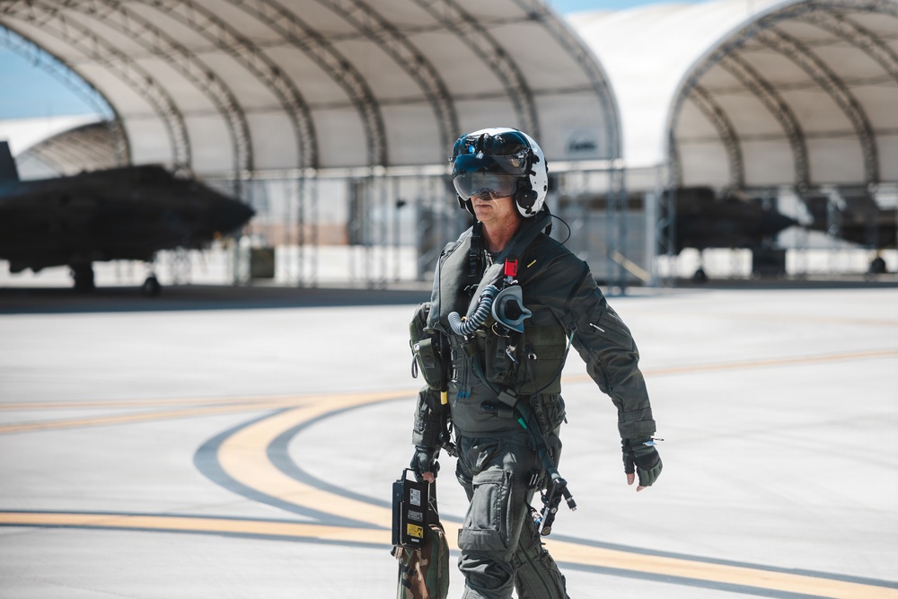 A military fighter pilot in full flight gear walks across an airfield tarmac toward the camera. The pilot is equipped with a modern flight helmet, oxygen mask assembly, G-suit, harness, and carries what appears to be a flight data or equipment case. In the background, arched aircraft shelters (HAS - Hardened Aircraft Shelters) house what appear to be dark-colored military jet aircraft.