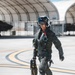 A military fighter pilot in full flight gear walks across an airfield tarmac toward the camera. The pilot is equipped with a modern flight helmet, oxygen mask assembly, G-suit, harness, and carries what appears to be a flight data or equipment case. In the background, arched aircraft shelters (HAS - Hardened Aircraft Shelters) house what appear to be dark-colored military jet aircraft.