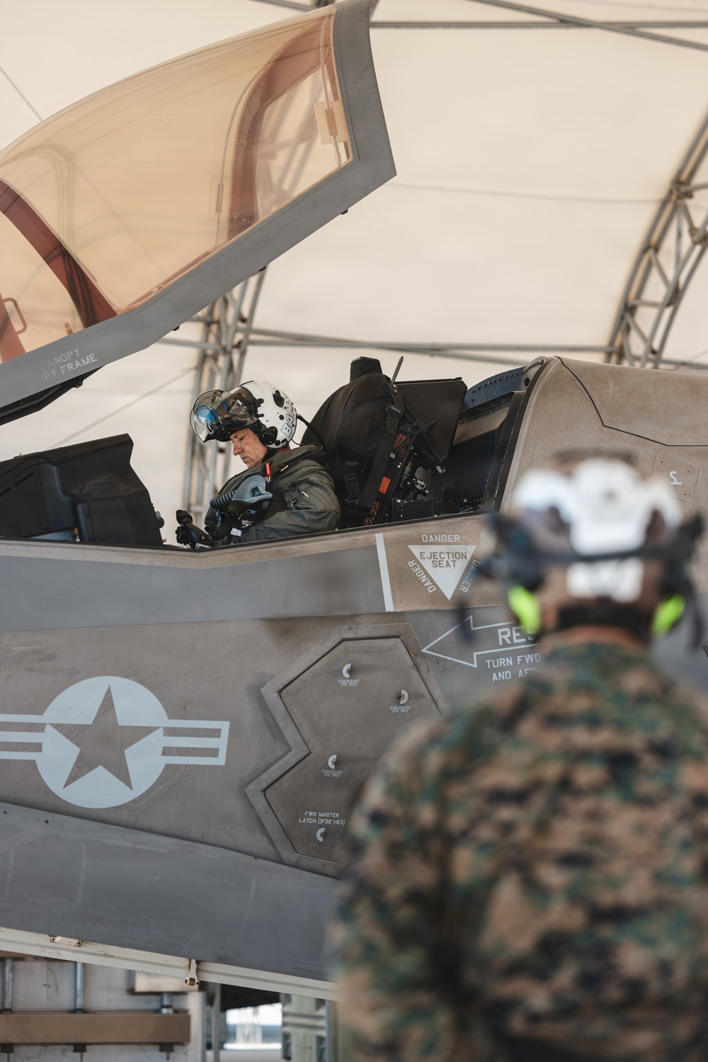 A military pilot is seated in the cockpit of an F-35 fighter jet, wearing a flight helmet with a visor and full flight gear. The aircraft is positioned under a shelter or hangar structure, with ground crew personnel in camouflage uniforms visible in the foreground. The image captures a pre-flight or post-flight operational scenario.