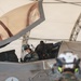 A military pilot is seated in the cockpit of an F-35 fighter jet, wearing a flight helmet with a visor and full flight gear. The aircraft is positioned under a shelter or hangar structure, with ground crew personnel in camouflage uniforms visible in the foreground. The image captures a pre-flight or post-flight operational scenario.