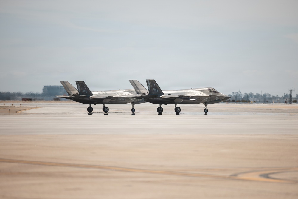 Two F-35 stealth fighter jets are taxiing side by side on a military airfield tarmac. Both aircraft display 'WF' tail codes, indicating they belong to the same unit. The image was taken under overcast skies with a flat, expansive concrete taxiway in the foreground.