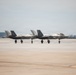 Two F-35 stealth fighter jets are taxiing side by side on a military airfield tarmac. Both aircraft display 'WF' tail codes, indicating they belong to the same unit. The image was taken under overcast skies with a flat, expansive concrete taxiway in the foreground.