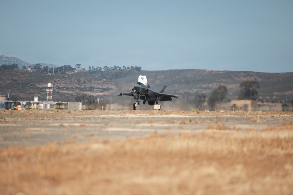 A military jet aircraft, consistent with an F-35 design, is captured during a takeoff or landing roll on a dry, arid airfield runway. The surrounding terrain features brown, drought-stressed vegetation and rolling hills in the background. Infrastructure elements including a radar dome and a red-and-white communications tower are visible to the left.