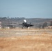 A military jet aircraft, consistent with an F-35 design, is captured during a takeoff or landing roll on a dry, arid airfield runway. The surrounding terrain features brown, drought-stressed vegetation and rolling hills in the background. Infrastructure elements including a radar dome and a red-and-white communications tower are visible to the left.
