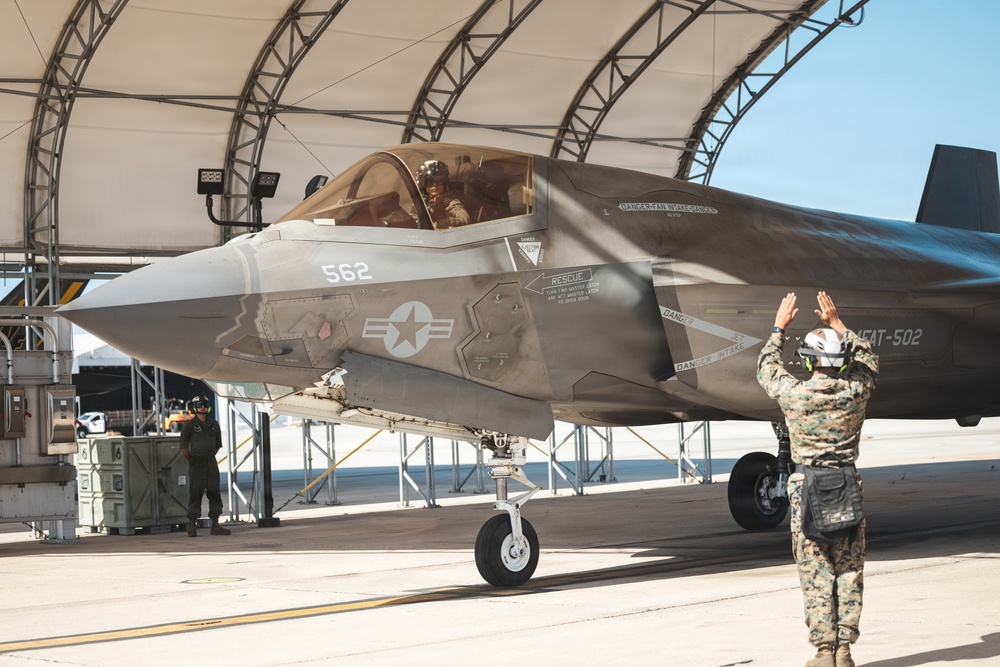 A U.S. Marine Corps F-35B Lightning II stealth fighter jet (bureau number 562) is being marshaled out of a hangar shelter by a ground crew member in camouflage utilities and a white helmet. A pilot is visible in the cockpit, and a second crew member stands near the hangar in the background. The scene appears to take place on a military air base flight line under clear skies.