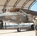 A U.S. Marine Corps F-35B Lightning II stealth fighter jet (bureau number 562) is being marshaled out of a hangar shelter by a ground crew member in camouflage utilities and a white helmet. A pilot is visible in the cockpit, and a second crew member stands near the hangar in the background. The scene appears to take place on a military air base flight line under clear skies.