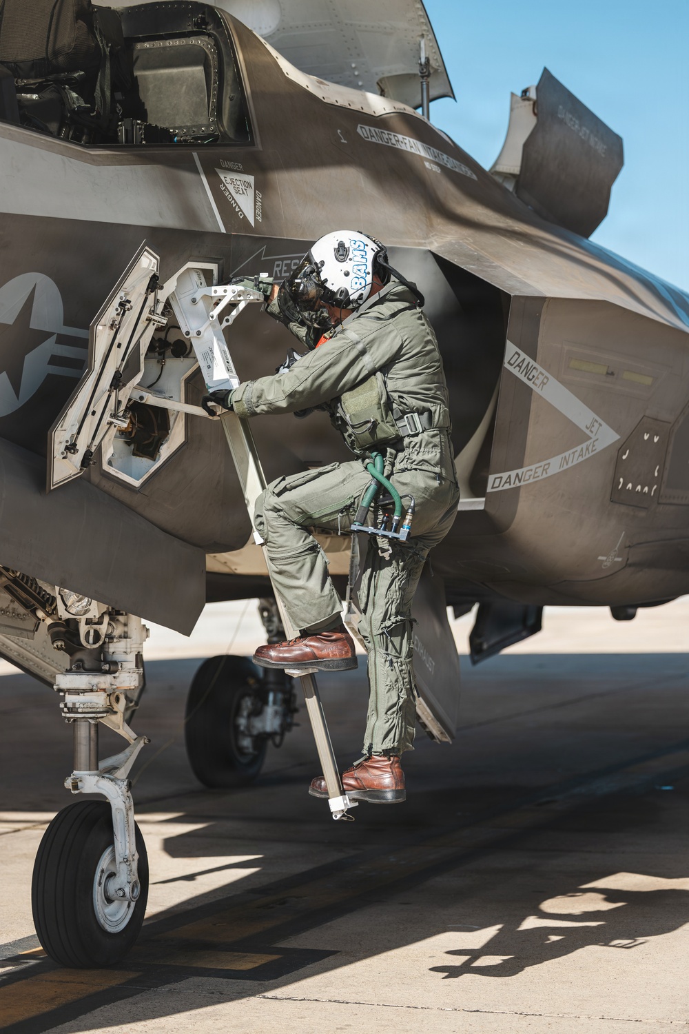 A military pilot in full flight gear is climbing into or performing a pre-flight inspection of what appears to be an F-35 Lightning II fighter jet on a tarmac. The pilot is wearing a green flight suit, white helmet with 'BAHS' written on it, oxygen mask, and brown flight boots. The aircraft displays multiple 'DANGER' warning labels including references to ejection seat, jet intake, and fan/wing areas.