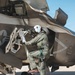 A military pilot in full flight gear is climbing into or performing a pre-flight inspection of what appears to be an F-35 Lightning II fighter jet on a tarmac. The pilot is wearing a green flight suit, white helmet with 'BAHS' written on it, oxygen mask, and brown flight boots. The aircraft displays multiple 'DANGER' warning labels including references to ejection seat, jet intake, and fan/wing areas.
